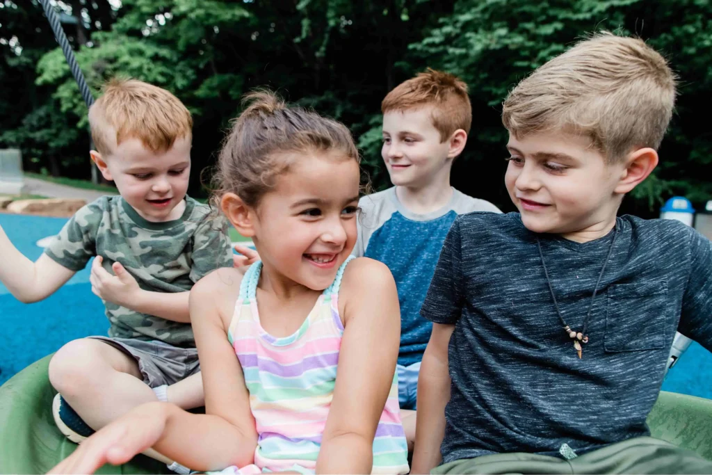 A group of four diverse, smiling children playing together on a green piece of playground equipment with a lush green forest background.