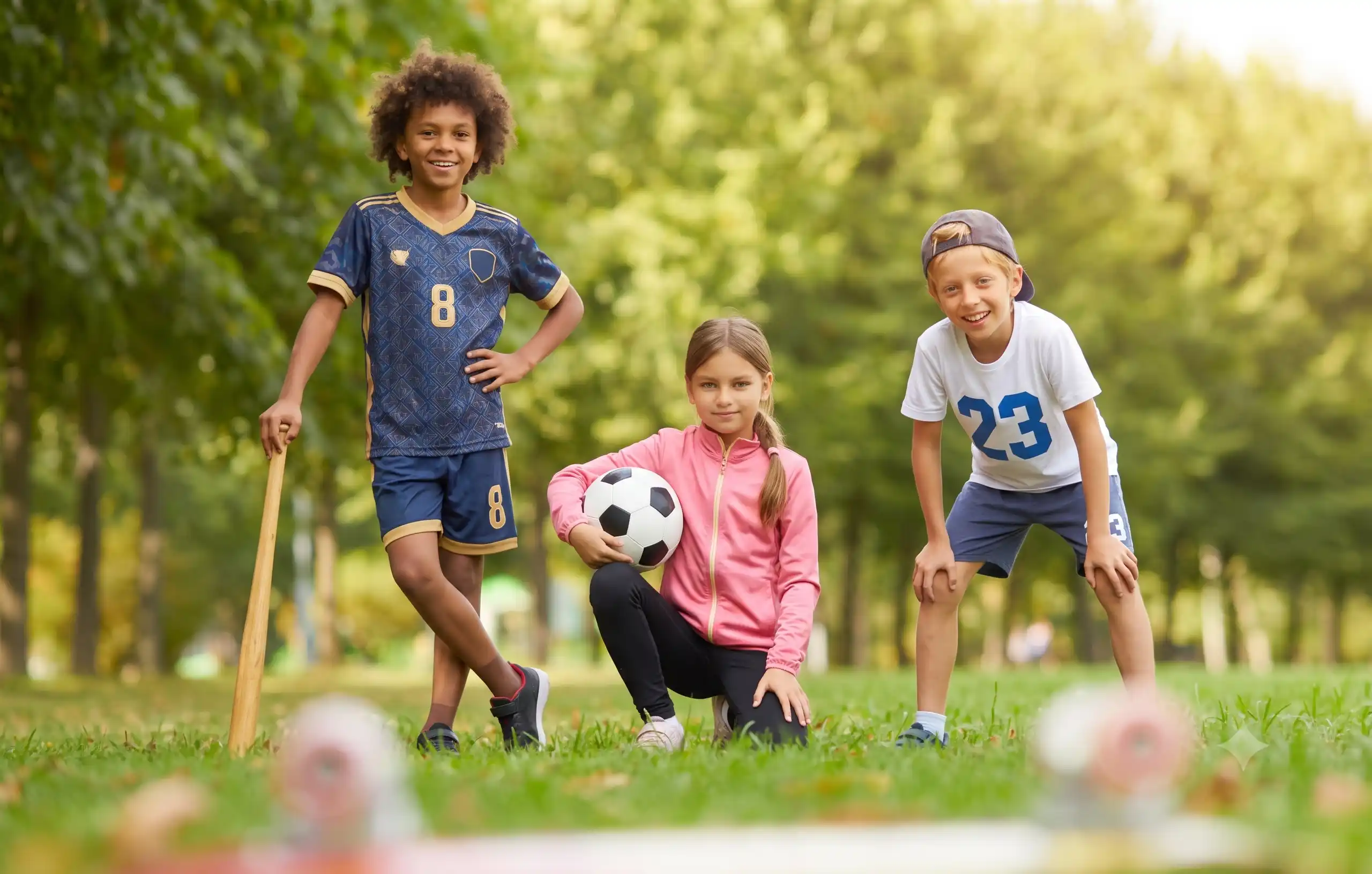 Three smiling children with sports equipment, including a soccer ball and a baseball bat, standing together in a grassy park.